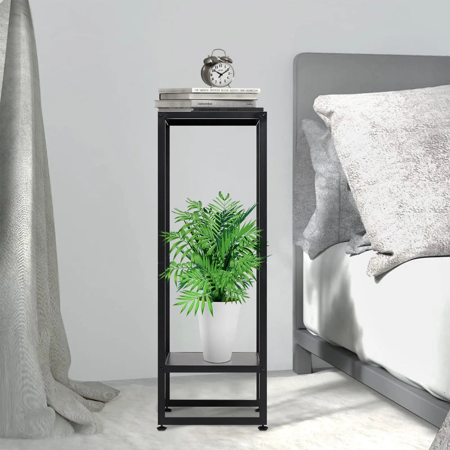 A black metal plant stand in a bedroom setting. The top level holds books and an alarm clock, while the bottom level features a white pot with a vibrant green plant. The stand is placed beside a bed with gray bedding.