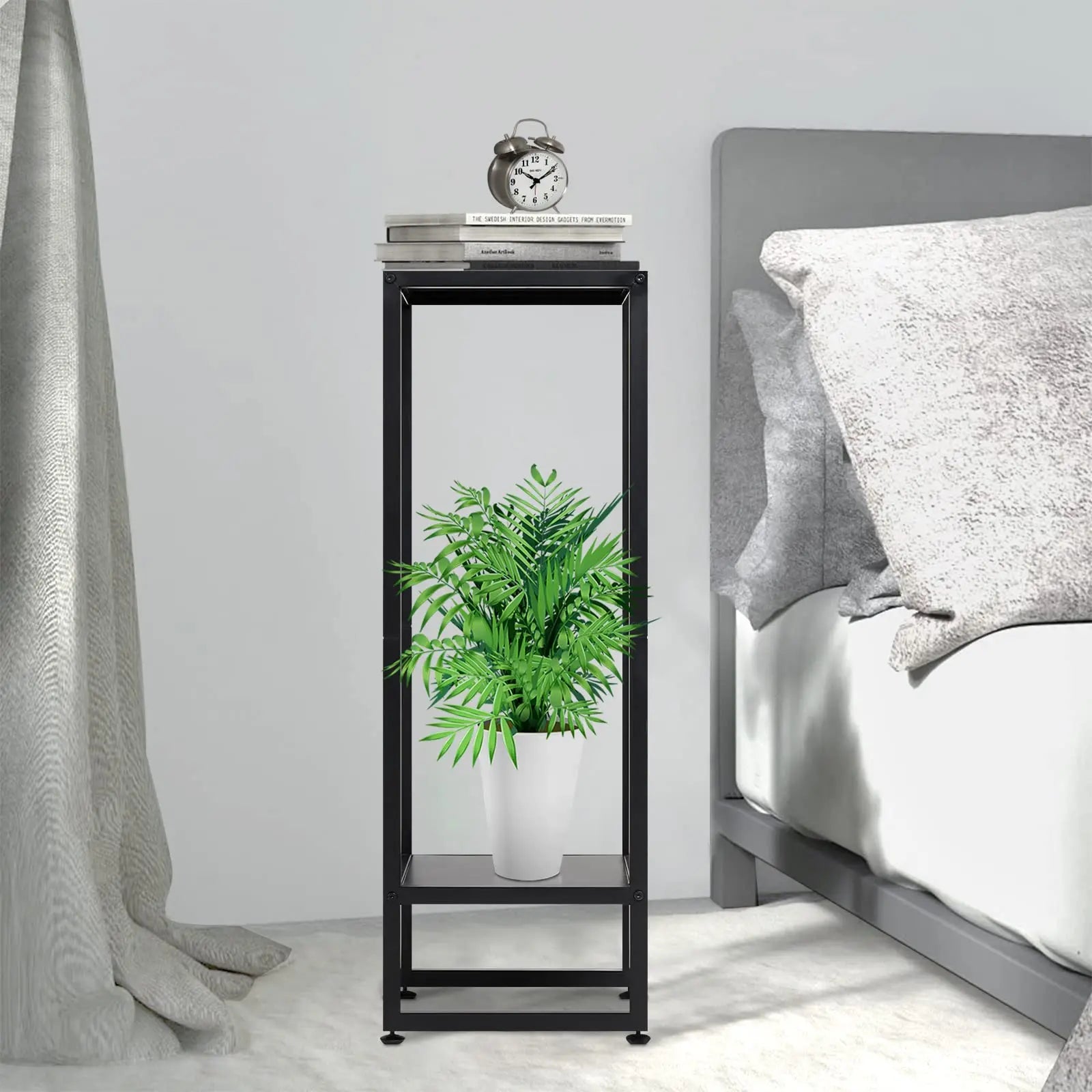  A black metal plant stand in a bedroom setting. The top level holds books and an alarm clock, while the bottom level features a white pot with a vibrant green plant. The stand is placed beside a bed with gray bedding.