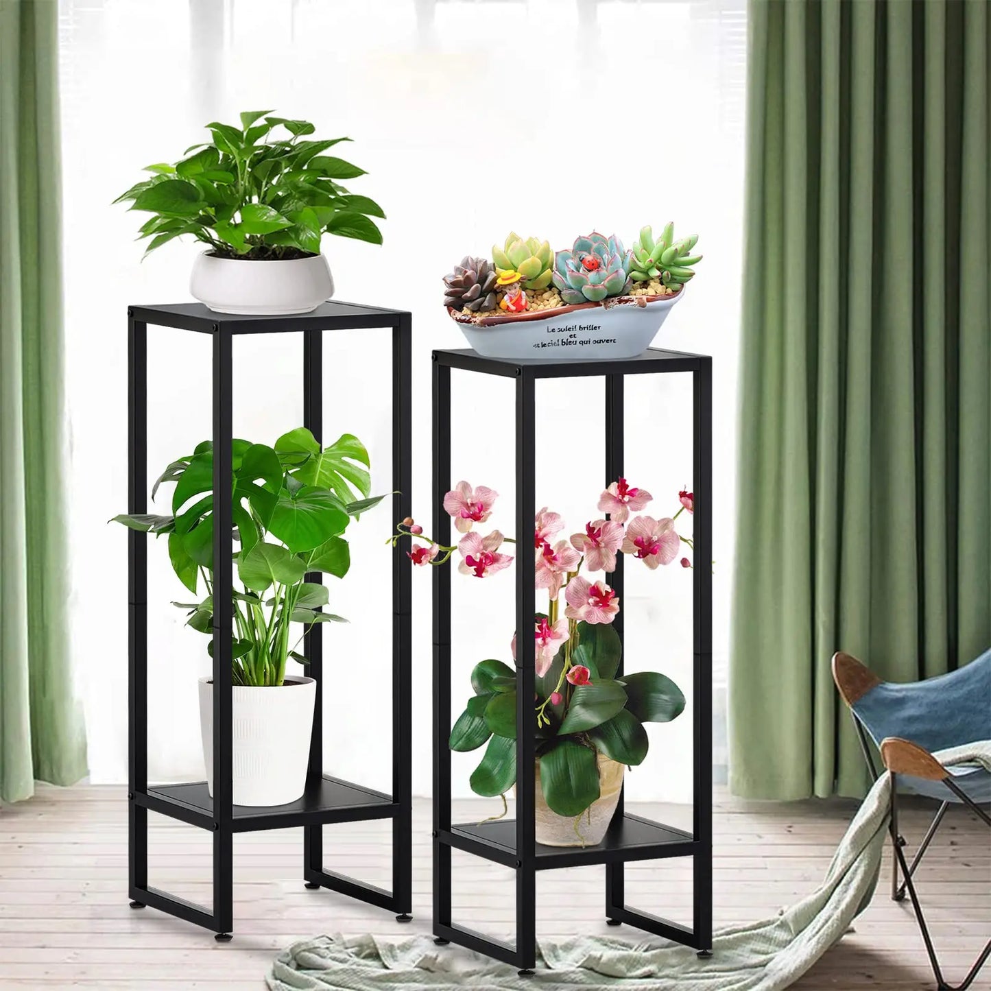 Two black metal plant stands in a room with green curtains. The left stand features a white pot with green leafy plants, and the right stand displays a white pot with pink orchids. The top of the right stand holds a decorative bowl of succulents.